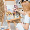 A blonde woman paints a miniature scene on a canvas using a detail paint brush set with natural wood handles.