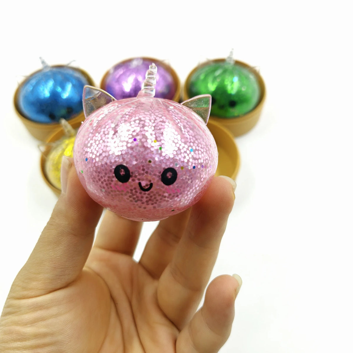 A hand holds a pink glitter unicorn dumpling squishy with a face, surrounded by other colored squishies.