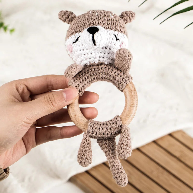 A hand holds an otter wooden baby rattle and teether ring made of knitted fabric on a white background.