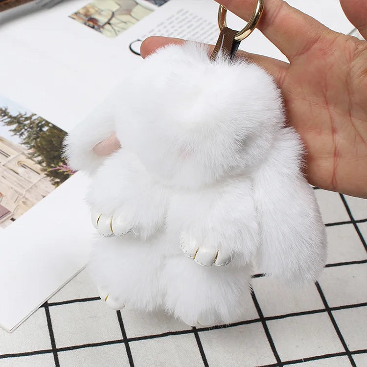 A fluffy white plush bunny keychain is being held by a hand, resting on a white surface.