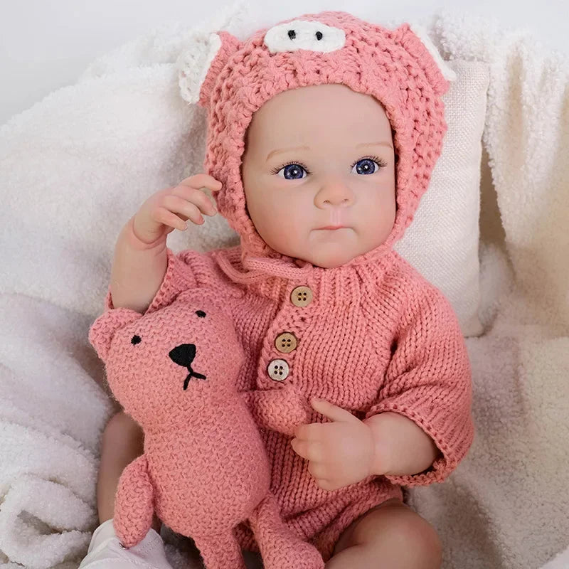 A realistic silicone reborn baby doll wearing a pink knitted outfit and a matching hood, resting in white fluffy fabric.