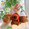 A hand holds a brown capybara plush keychain charm with a green leaf and a white knitted hat.