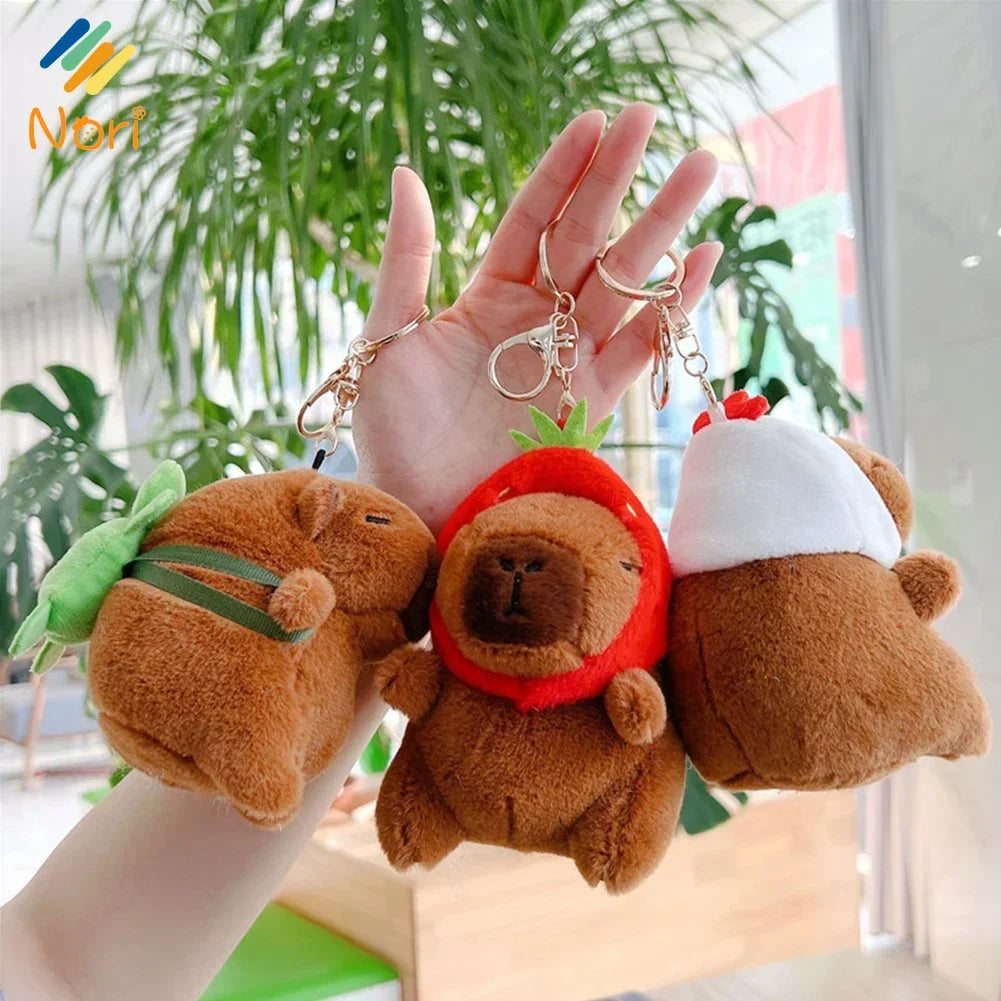 A hand holds a brown capybara plush keychain charm with a green leaf and a white knitted hat.
