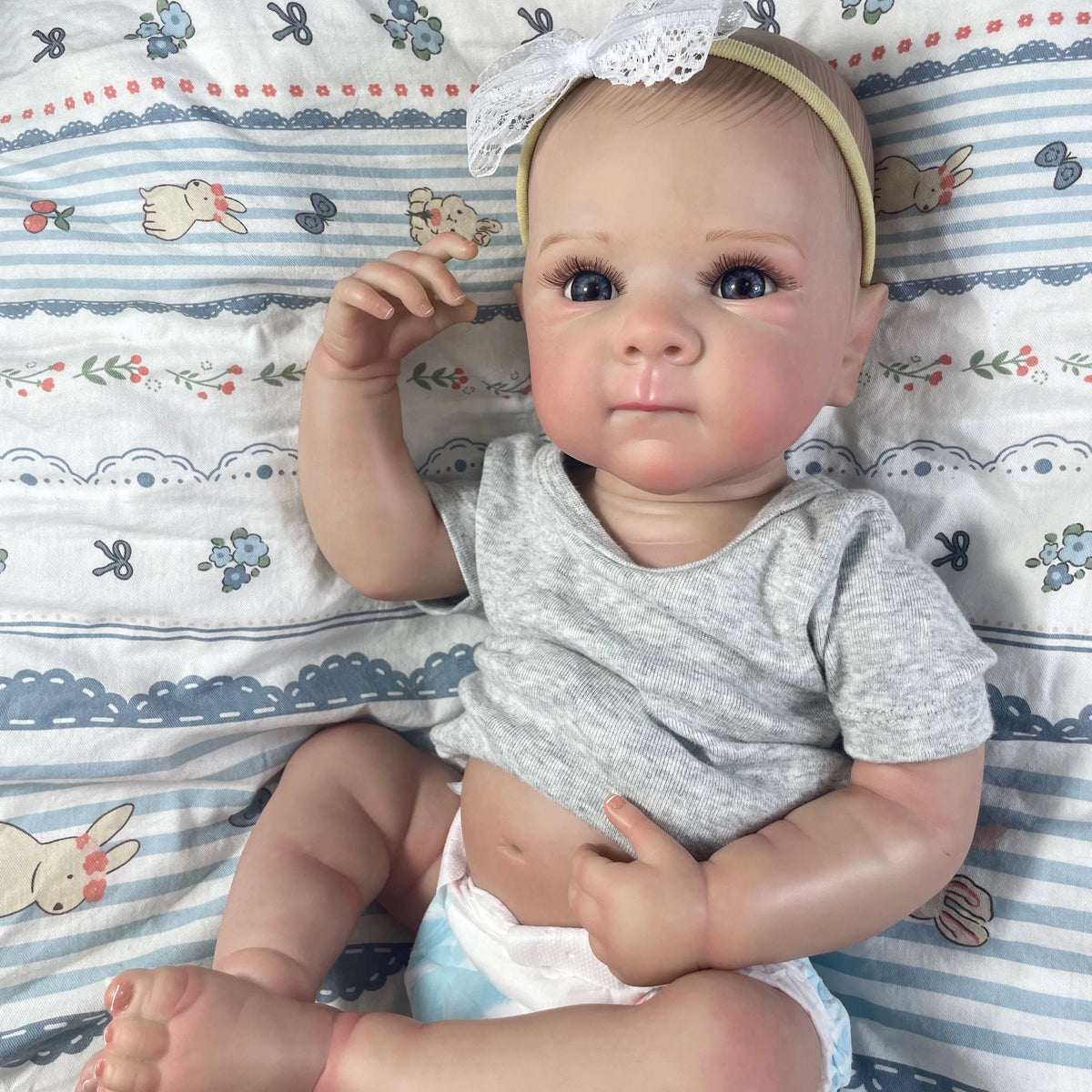 A realistic reborn baby doll with painted hair wearing a gray shirt, lying on a striped blanket.
