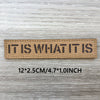 Un patch tissé marron rectangulaire avec l'inscription IT IS WHAT IT IS, mesurant 12x2.5 cm.