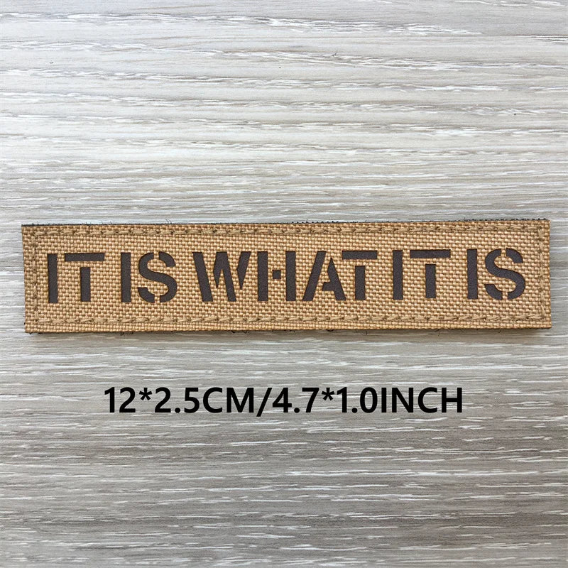 Un patch tissé marron rectangulaire avec l'inscription IT IS WHAT IT IS, mesurant 12x2.5 cm.