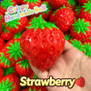 A hand holds a bright red strawberry stress ball with green leaves, surrounded by other similar toys.