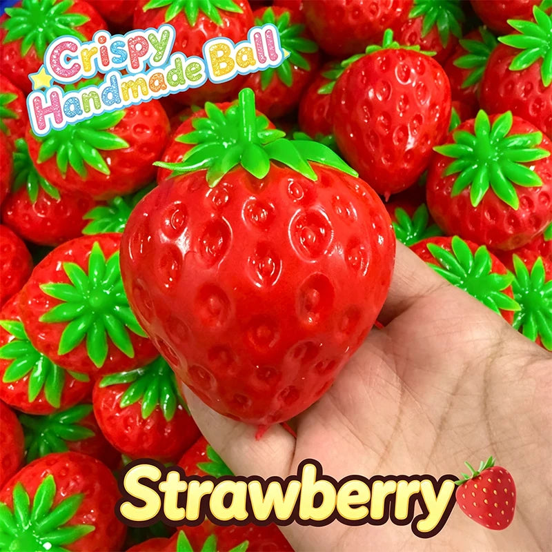 A hand holds a bright red strawberry stress ball with green leaves, surrounded by other similar toys.
