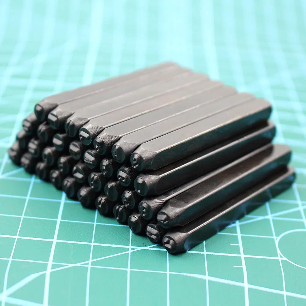 A stack of 36 carbon steel number and letter stamping rods resting on a teal grid surface.