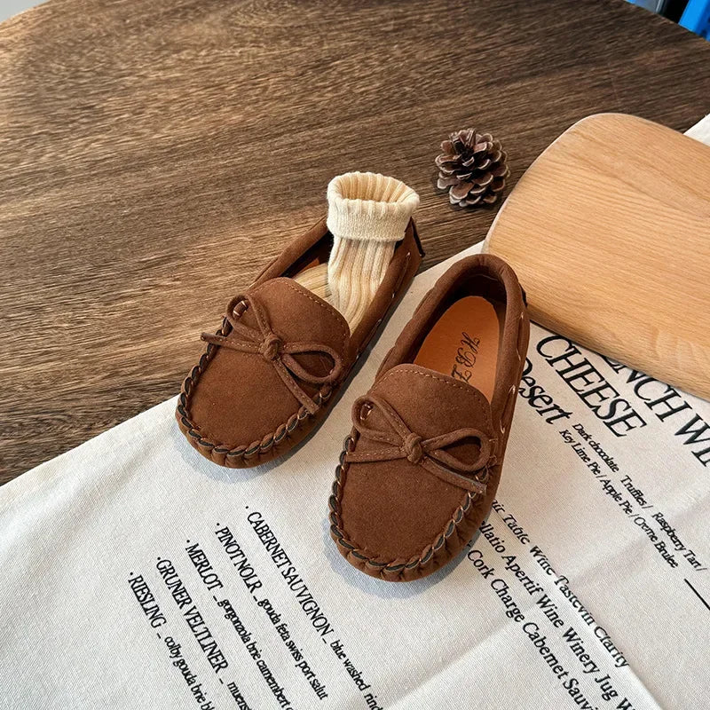 A pair of brown girls' moccasins with bow details resting on a white paper.