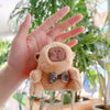 A hand holds a small, fluffy brown capybara plush keychain charm against a blurred green background.
