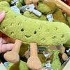 A soft green plush pickle toy with a smiling face and brown legs is shown in a box.
