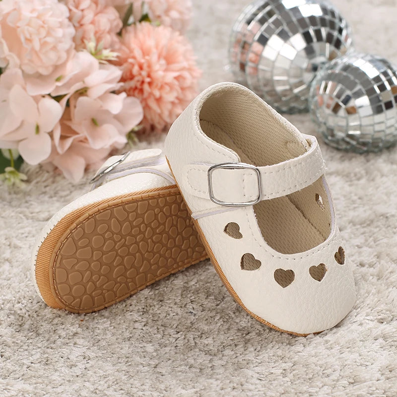 A pair of cream-colored moccasin shoes with lace trim and heart cutouts resting on a fuzzy surface next to pink flowers and s