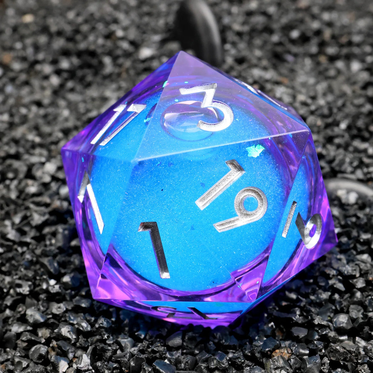 A handmade resin D20 die with a large blue and purple liquid swirl, viewed from above on black gravel.