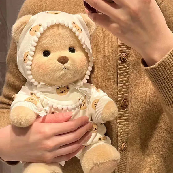 A cute plush teddy bear doll wearing a white outfit with bear patterns, held by a person against a beige sweater.