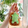 A hand holds an adorable capybara plush keychain charm with green skin and brown fur against a blurred indoor background.