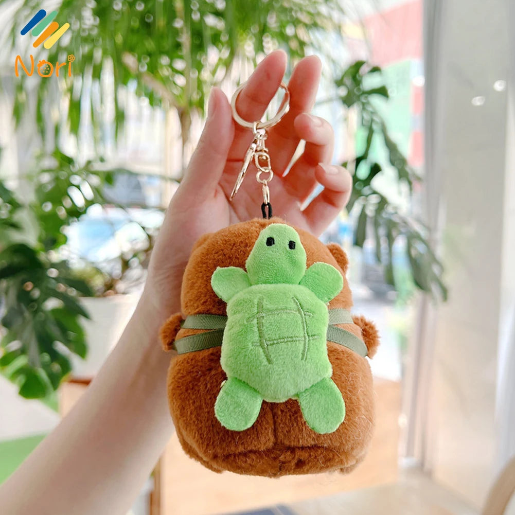 A hand holds an adorable capybara plush keychain charm with green skin and brown fur against a blurred indoor background.