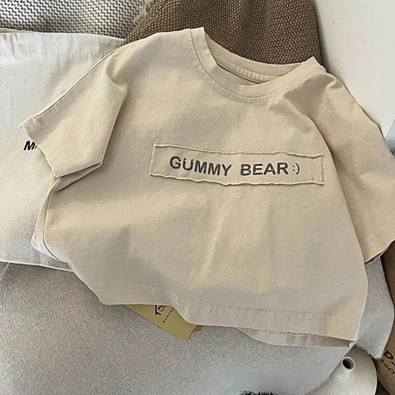 A light beige short-sleeve t-shirt featuring embroidered text that reads GUMMY BEAR.