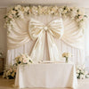 Oversized creamy white satin bow decoration draped across a white backdrop for an event or party setting.