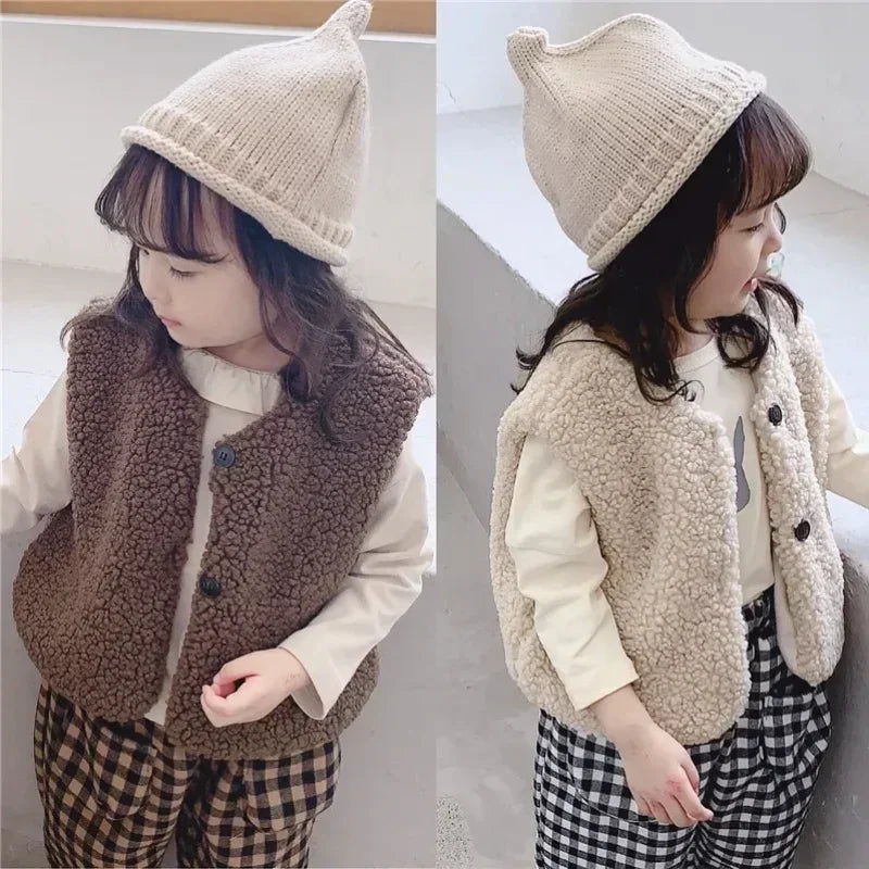 A young girl wearing a beige plush fleece vest over a white shirt and plaid pants.