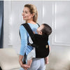 A woman wearing a blue shirt is holding a baby in an ergonomic black baby carrier with adjustable straps.