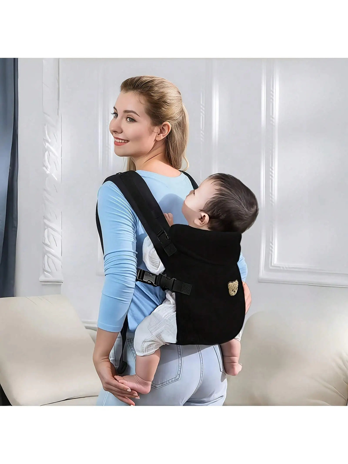 A woman wearing a blue shirt is holding a baby in an ergonomic black baby carrier with adjustable straps.