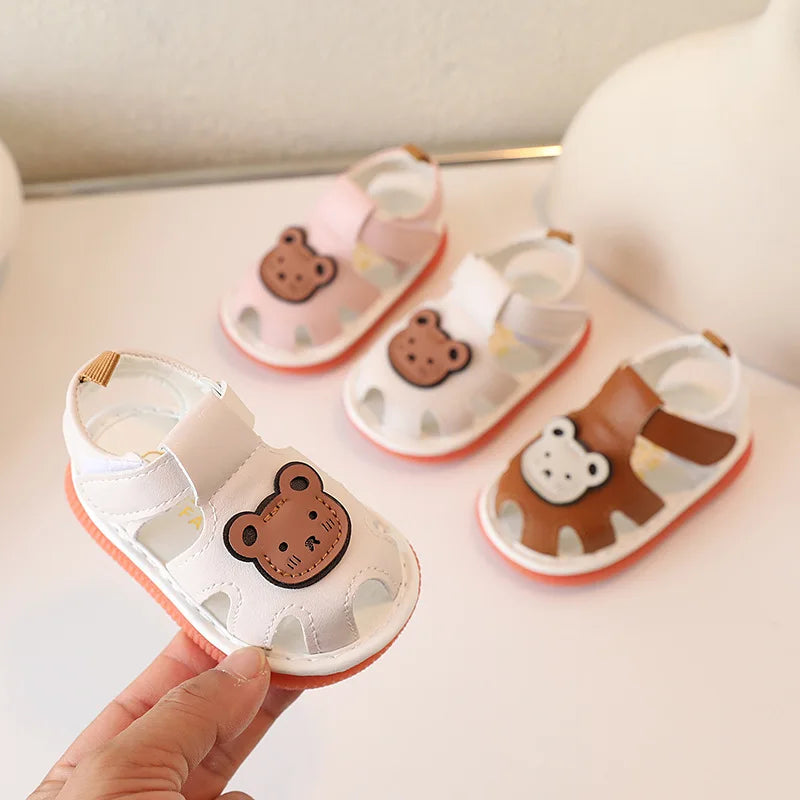 A hand holds a pair of cute bear soft sole sandals for infants and toddlers, showing various colors and designs.
