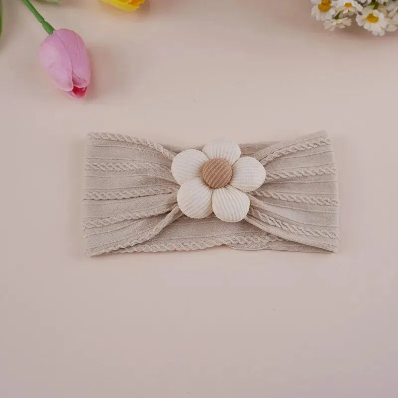A beige knitted elastic headband with a white flower detail is shown on a light pink background.