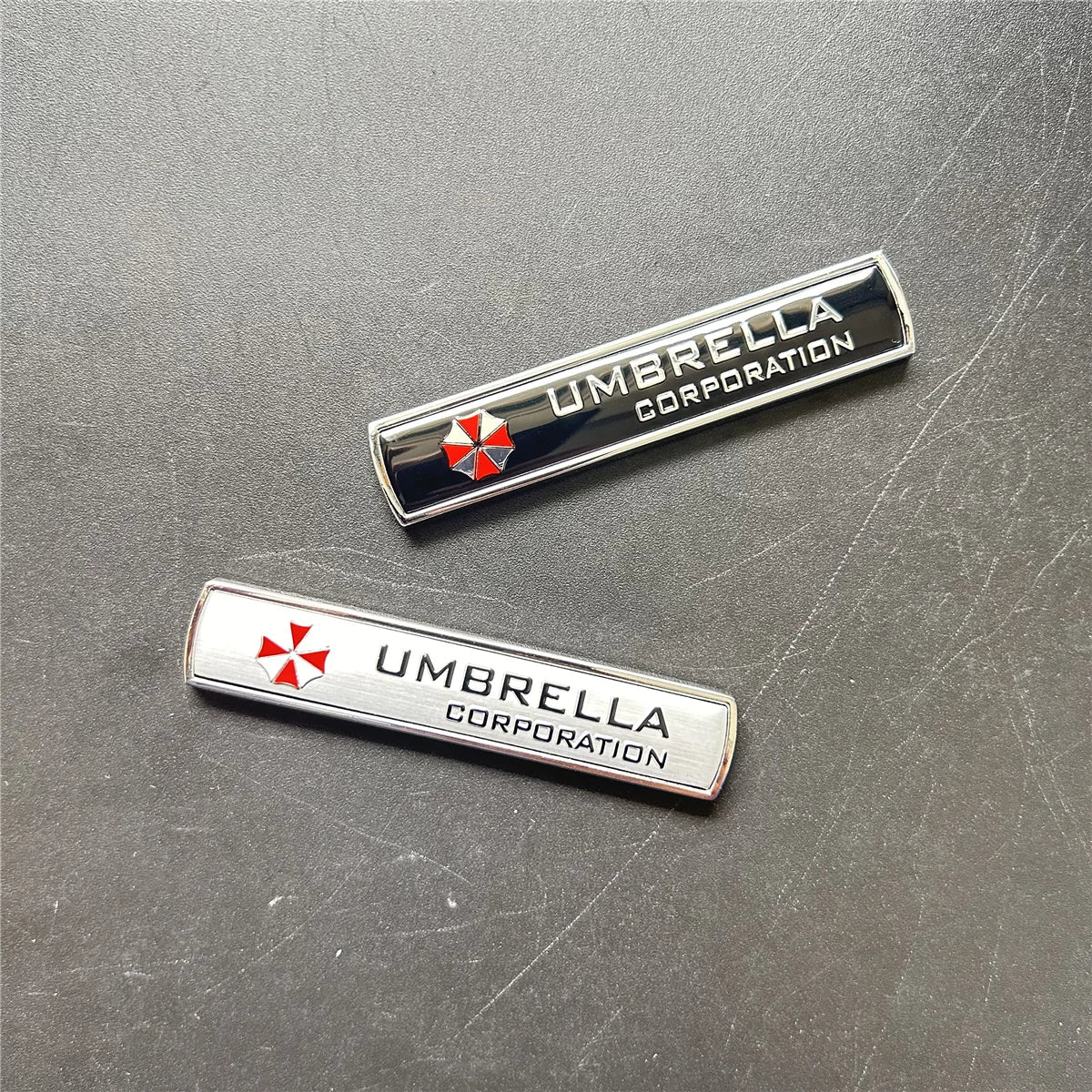 Two silver metal badges featuring the Umbrella Corporation logo and name on a dark gray surface.