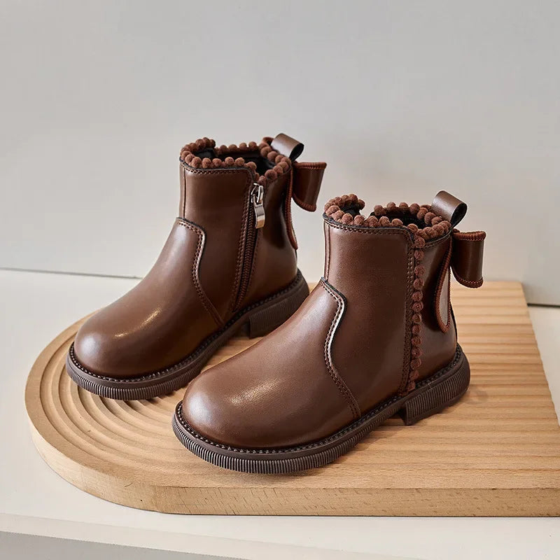 A pair of brown leather ankle boots for girls featuring a bow-knot detail and a side zipper, resting on a wooden surface.