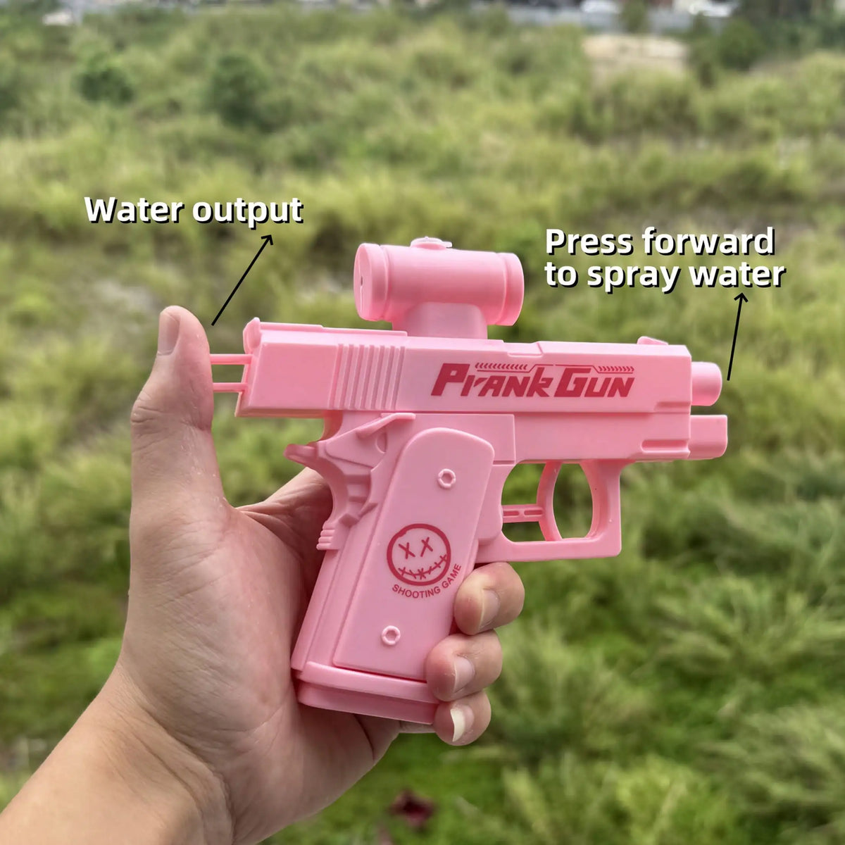 A hand holds a pink prank gun water toy, demonstrating the water output feature in an outdoor setting.
