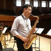 A man plays a brass saxophone on a stage with wooden seating and windows in the background.