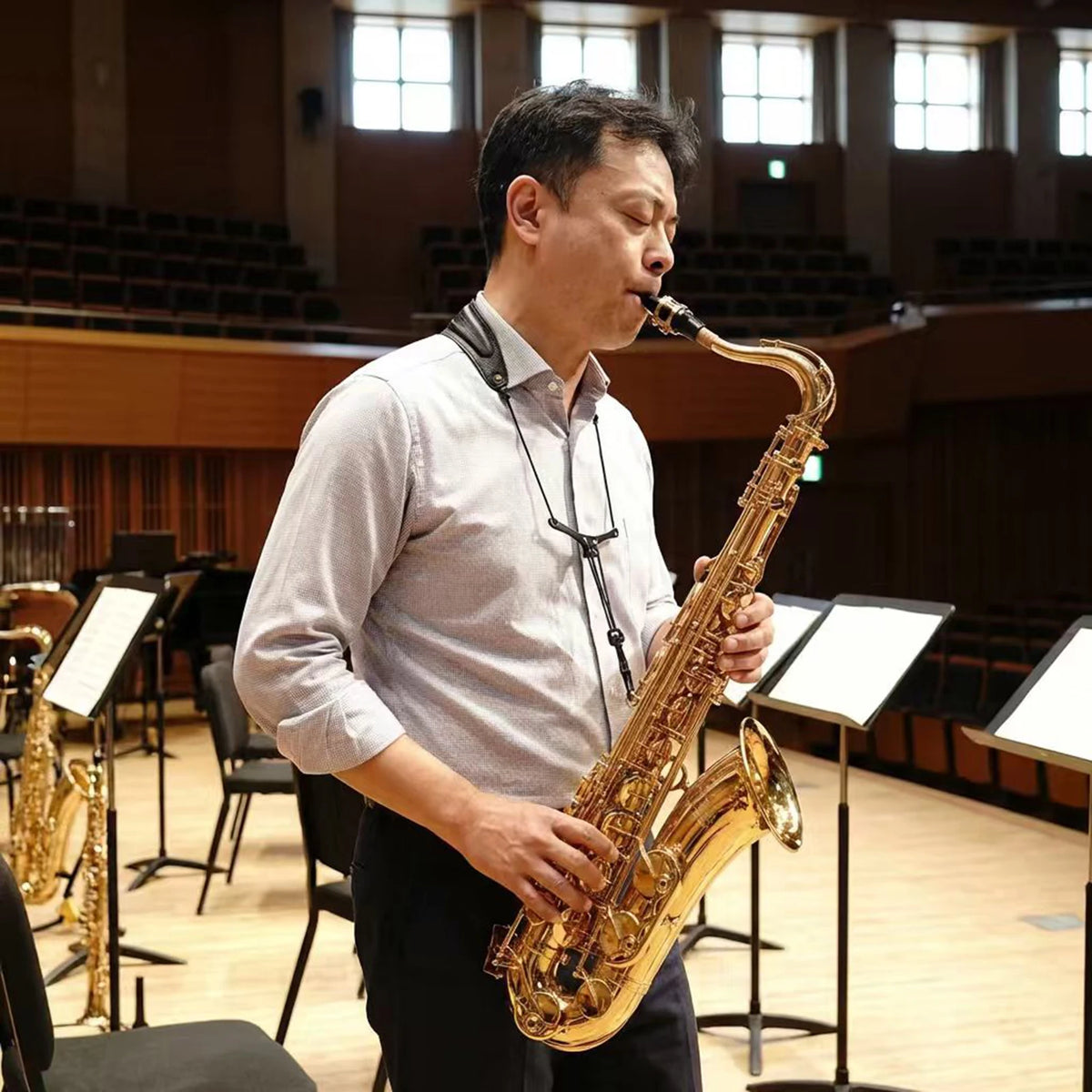 A man plays a brass saxophone on a stage with wooden seating and windows in the background.