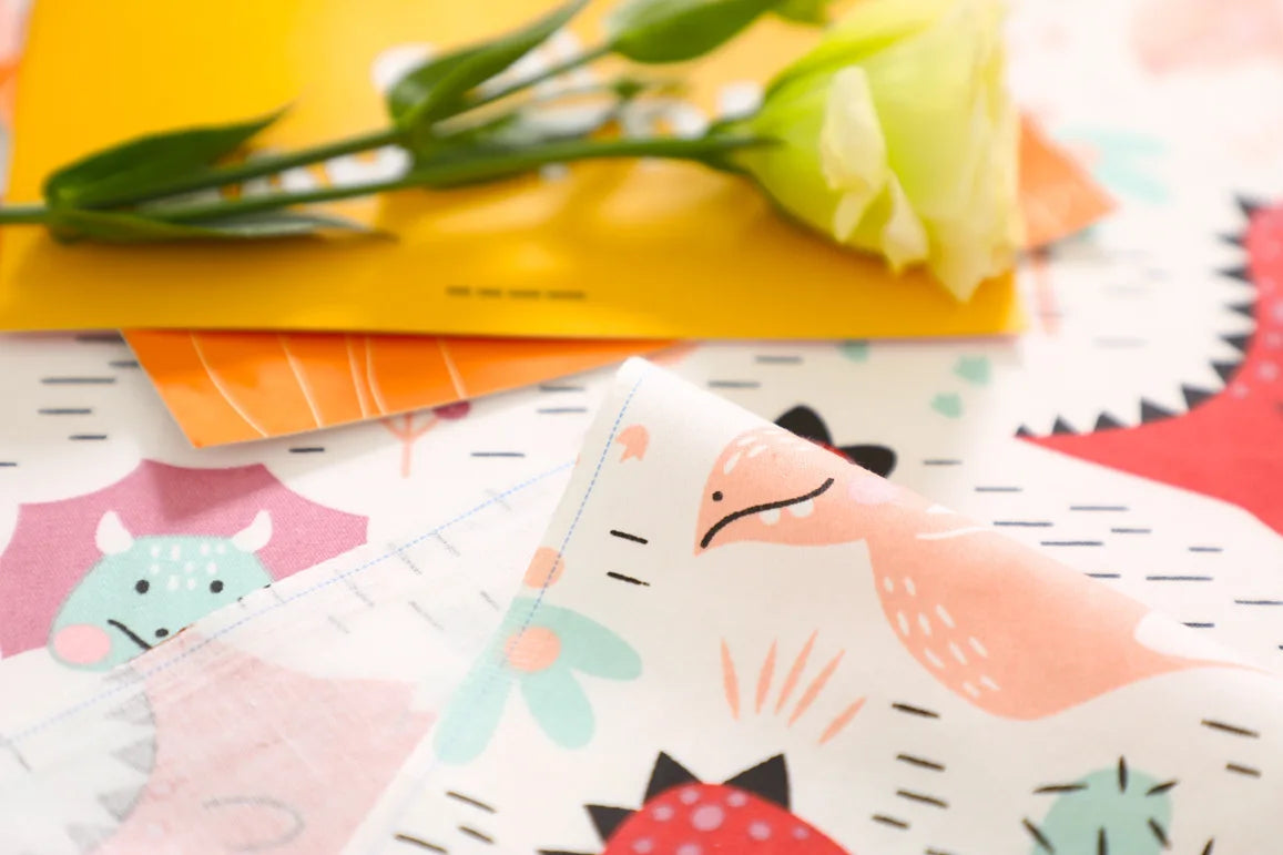 A close-up of a fabric with a dinosaur grid print featuring pink, green, and orange elements.
