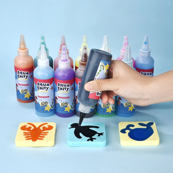 A hand is holding a bottle of Aqua Fairy liquid next to various colored bottles and wooden craft pieces.