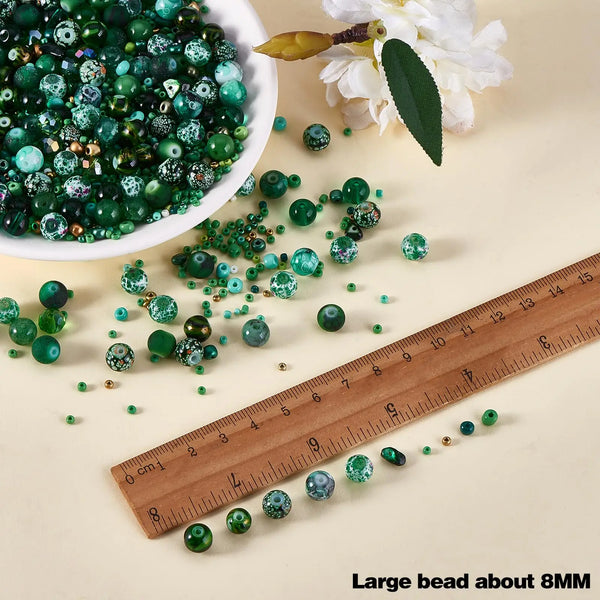 A collection of mixed size green glass seed beads scattered on a light background next to a ruler and white flower.