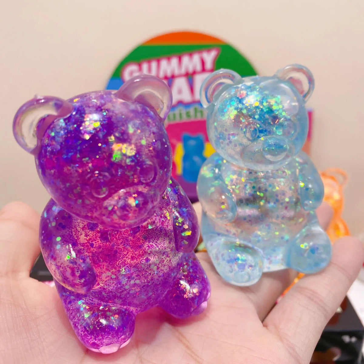A hand holds a purple glitter bear squishy toy next to a blue glitter bear squishy toy.