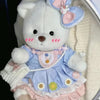 A white teddy bear plush wearing a light blue and pink checkered dress with a frilly collar.