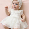 A baby girl wearing a white lace tulle dress and matching hat, looking up with blue eyes.