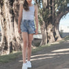 A young girl wearing a light gray tank top and denim shorts stands on a path with barefoot sneakers.