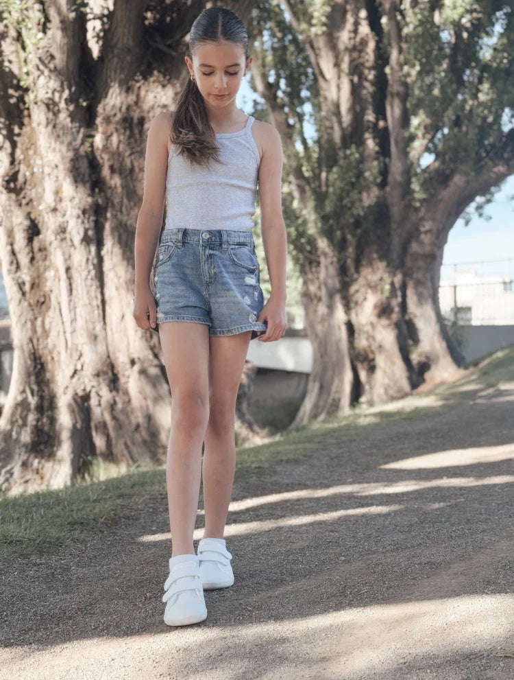 A young girl wearing a light gray tank top and denim shorts stands on a path with barefoot sneakers.