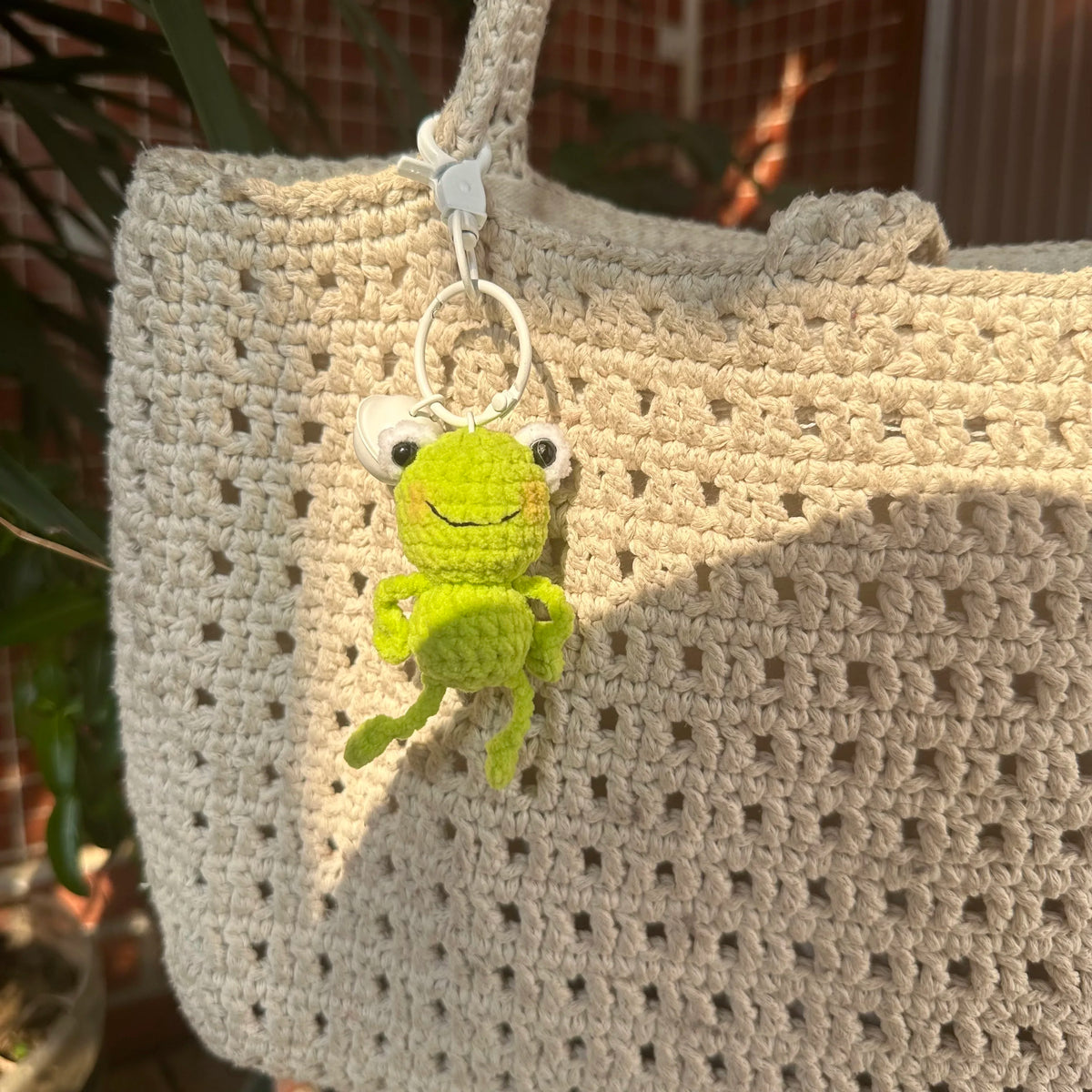 A handmade crochet frog keychain with bright green skin hanging from a light beige knitted bag.