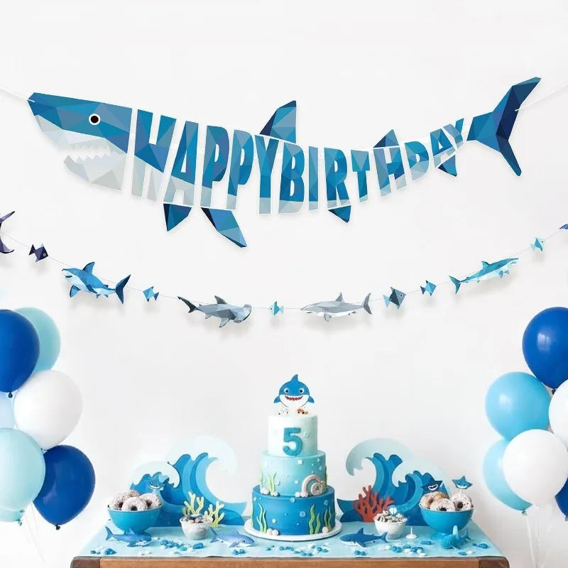 A blue geometric shark happy birthday banner and hanging garland set displayed on a wooden surface.