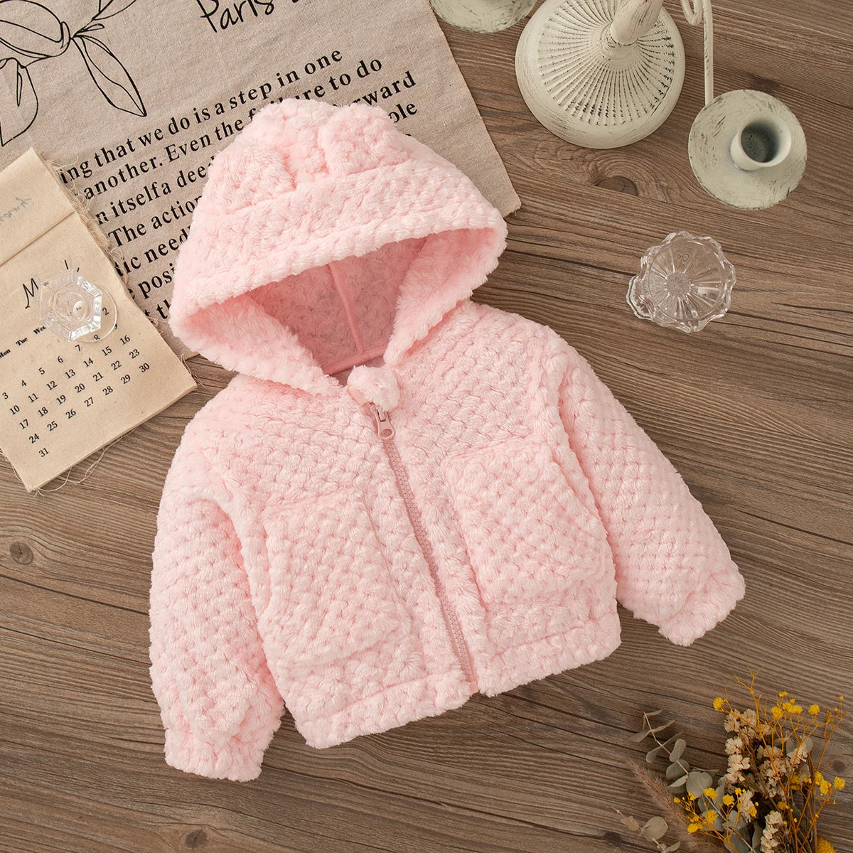 A pink plush hooded winter jacket for a baby girl lying on a wooden surface next to a calendar and decorative items.