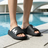 A pair of black Cloud Comfort EVA slide slippers with non-slip soles resting on a stone patio beside a swimming pool.