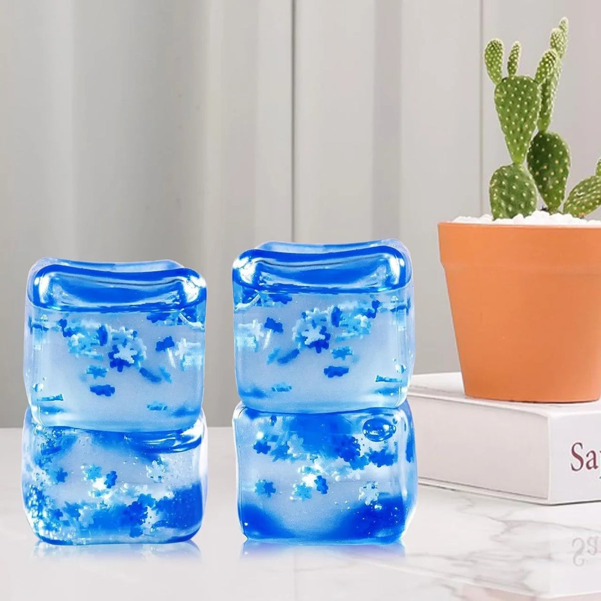 Two translucent blue squishy stress cubes with sparkling blue glitter are displayed next to a potted cactus.