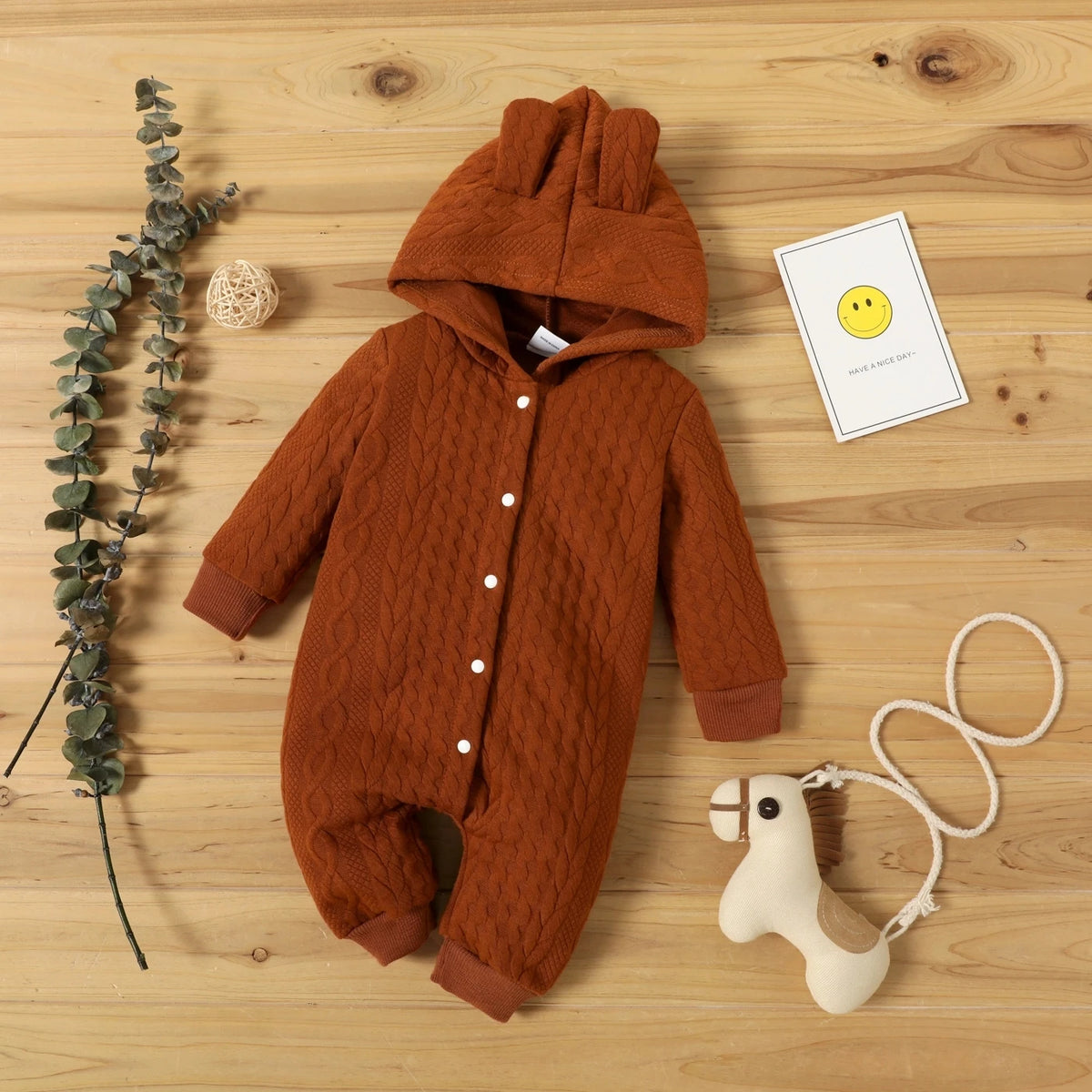 A brown knitted hooded baby romper lying on a wooden surface with a sprig of greenery and a yellow smiley card.