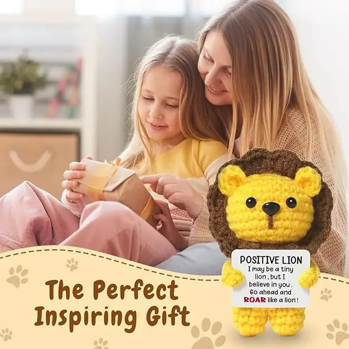 A young girl is holding a yellow crochet lion doll with a positive message card, being comforted by an adult.