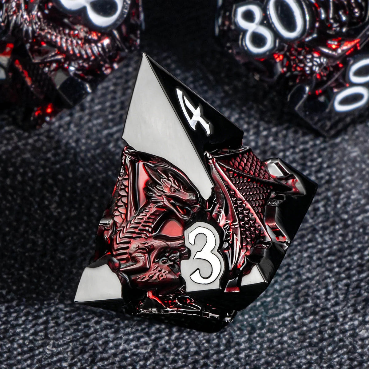 A close-up of a black and silver metal die featuring a dragon design and the number 3, set against a dark gray background.