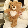 A brown teddy bear with a white heart patch is held by a person wearing a cream-colored sweater.
